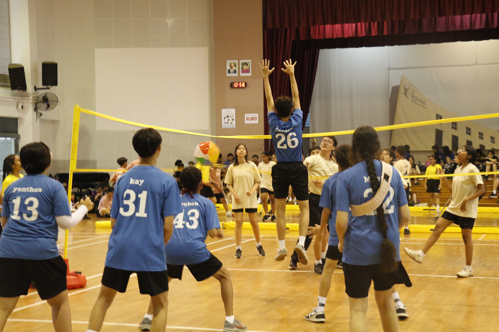 Students in blue and cream shirts play a volleyball match with a colorful beach ball inside a school sports hall.